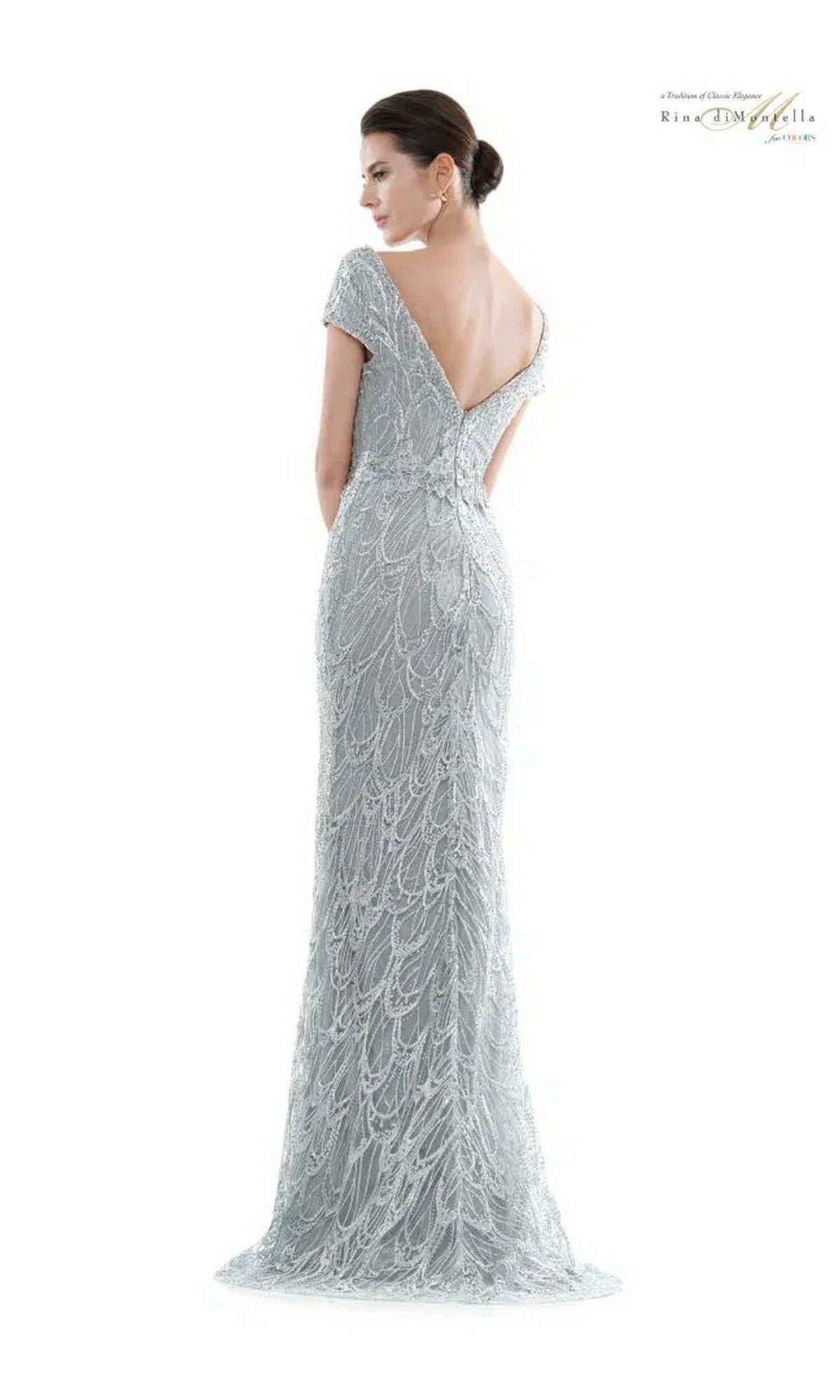 A woman stands facing away in the Long Formal Dress: Rina Di Montella RD2716, a light grey, floor-length gown with a mermaid silhouette and low V-back. Her hair is styled in an elegant updo.